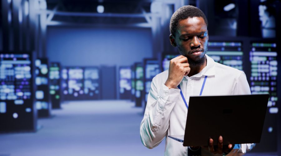 Electrician inspecting blade servers delivering online web content to customers. African american wireman using laptop to take note of critical systems, looking for damaging errors